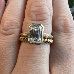 Shown on hand: emerald cut bezel engagement ring with hand engraved yellow gold band, stacked with a twist gold band