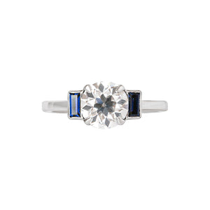 White gold engagement ring with four claw prongs and vertical lab grown sapphire baguettes, set with an Old European Cut moissanite or lab diamond.
