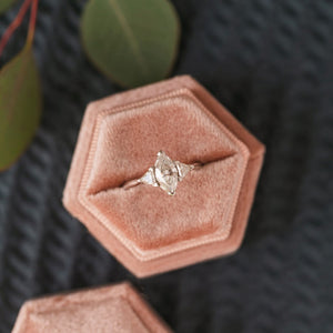 Three-stone marquise and trillion engagement ring in a pink ring box on a dark background.