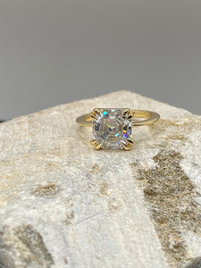 Asscher cut yellow gold engagement ring with double claw prongs rests on a piece of raw stone. Gray background.